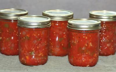 Five jars of canned stewed tomatoes sitting on a gray kitchen towel with a gray background.