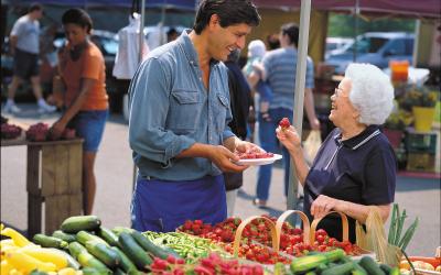 A male vendor serving a strawberry sample to an older woman at a farmers market stand.