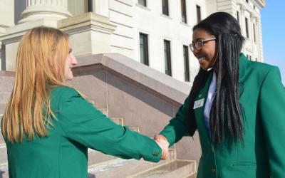 two ladies in green jackets shake hands in front of a building.