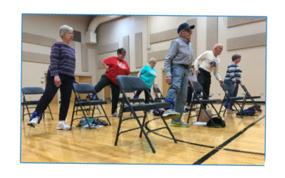 a group of older adults starting to exercise while standing behind folding chairs