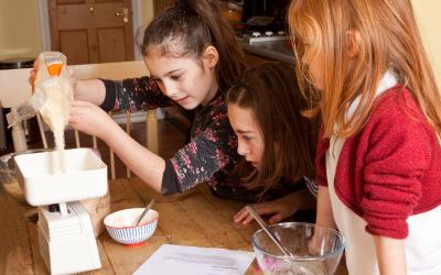 Three young girls weighing sugar on a kitchen scale.