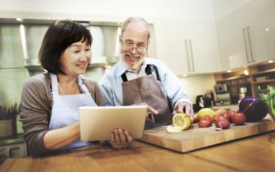 An older couple standing in the kitchen looking at a tablet while cutting a lemon on a wood cutting board