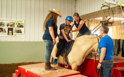 Staff of the Horse Power organization helping a young male aboard a horse for a therapeutic ride.