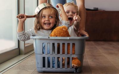 Mother pushing laundry basket along the floor with two children riding inside it.