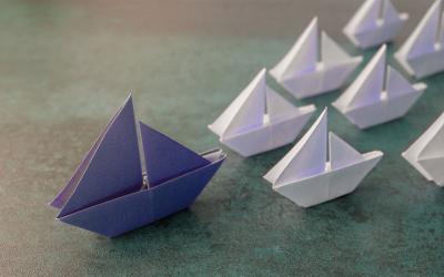 Purple origami paper sailboats leading several smaller, white paper sailboats.