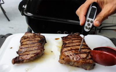 Hand holding meat thermometer to check the doneness of grilled steaks.