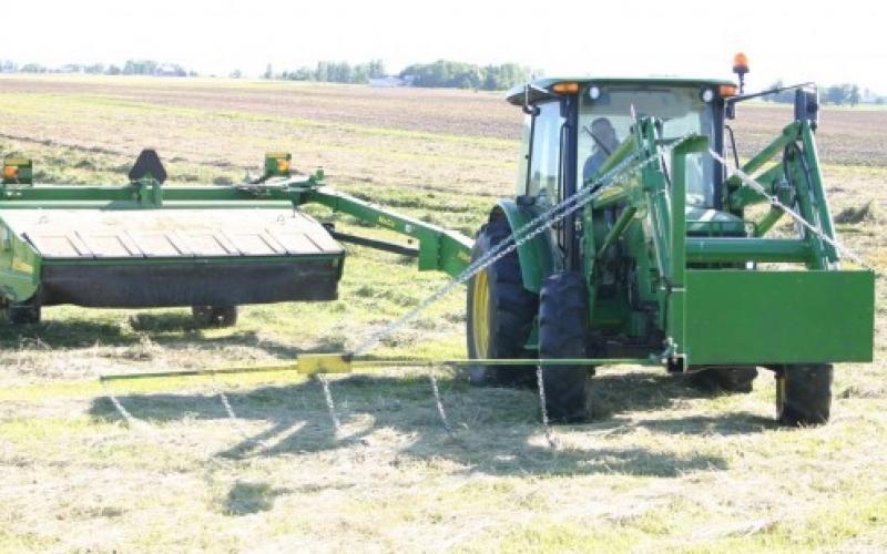A green front-end-loader pulling a hay mower with a flushing bar.
