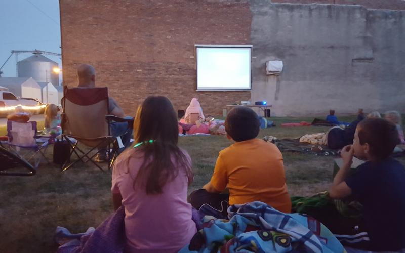 A group of people watching a movie outdoors on a video screen on the side of a brick building.