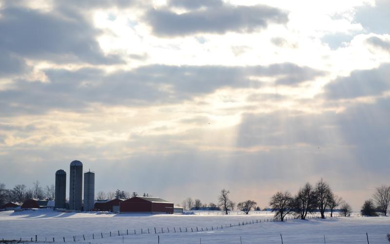 A snowy farmyard with sunlight breaking through the clouds.