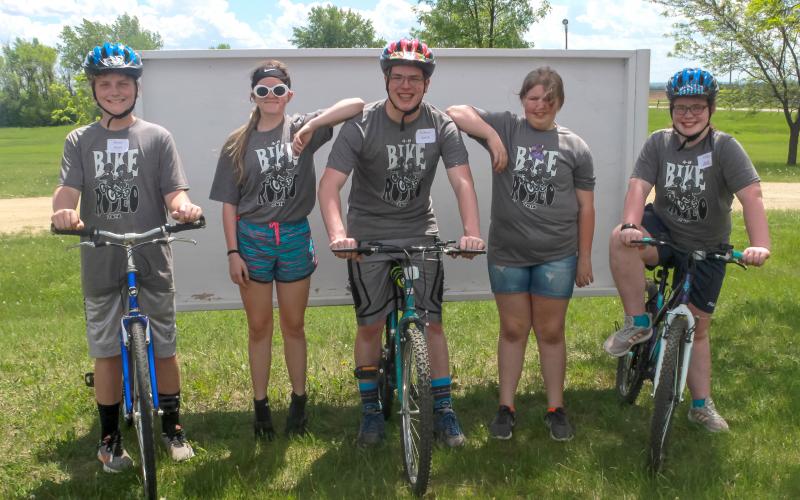 a group of five 4-H youth. two males and one female are on bicycles. two female youth are standing in the background.