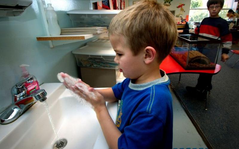 young boy washing hands at a sink in a classroom.