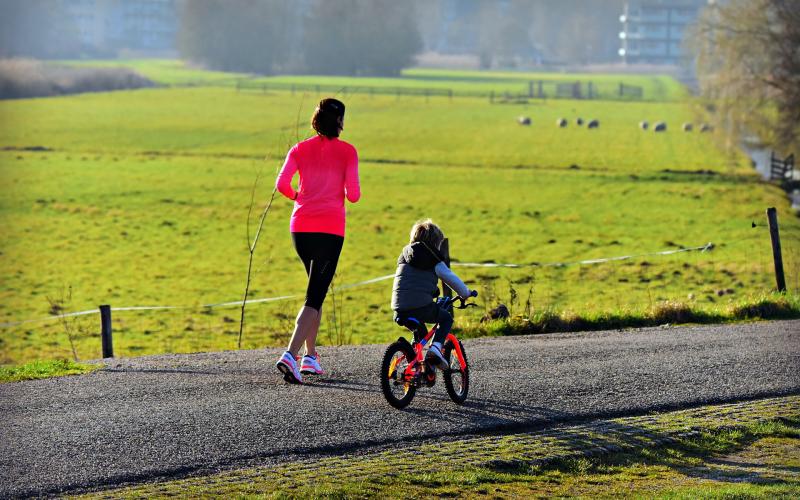 A mother jogging down a paved country trail next to a child riding an orange bicycle.