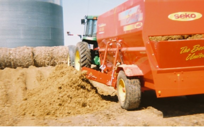 A green tractor pulling a red wagon next to a pile of wet distillers grains.