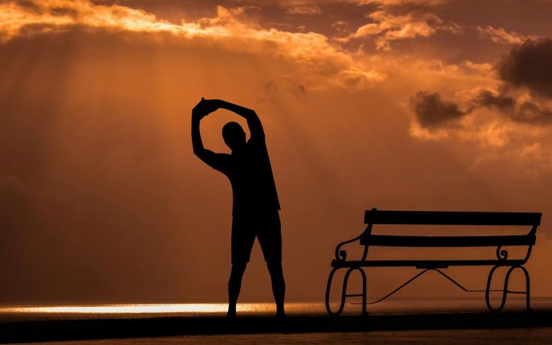 person stretching in front of the rising sun near a bench on the beach