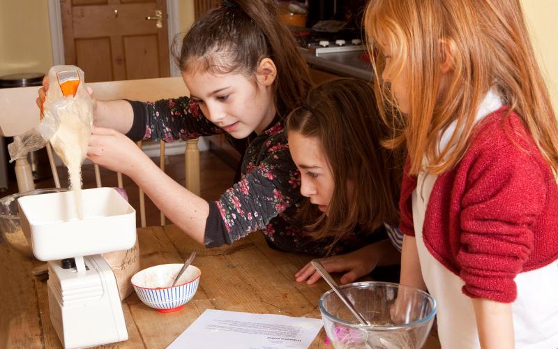 Three young girls weighing sugar on a kitchen scale.