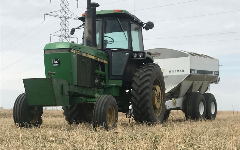 A green tractor pulling a fertilizer wagon through a field of alfalfa.