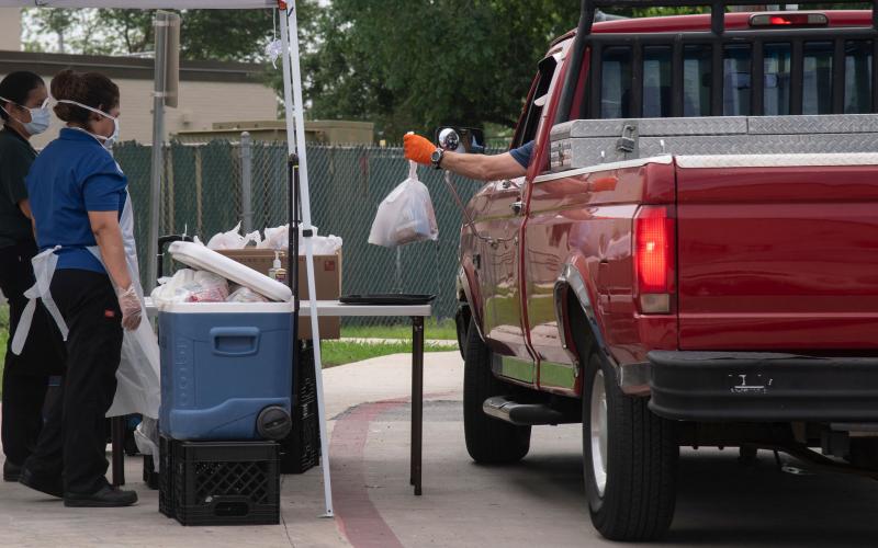 A man in a pickup truck picking up a bag of farmers market food at a pickup table being serviced by two women in personal protective gear.