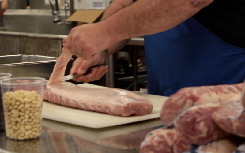 A man in a blue apron trimming the fat off the back of a pork loin cut.