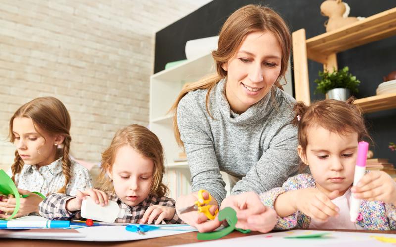 A mother with three children at a craft table cutting and gluing pieces of colored paper.