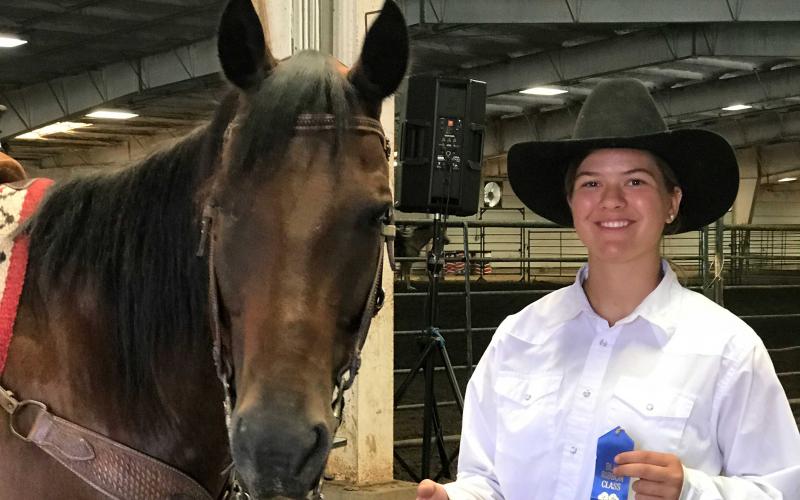 Rose Eitemiller holding a blue ribbon. She is standing next her horse in an arena.