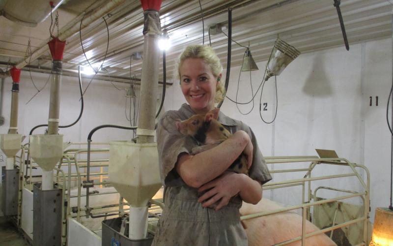 A woman standing inside a pig barn and holding a piglet