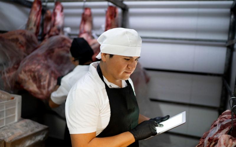 Young producer inspecting meat in a locker with a digital tablet.