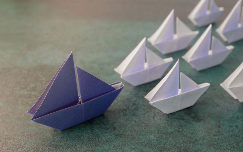 Purple origami paper sailboats leading several smaller, white paper sailboats.