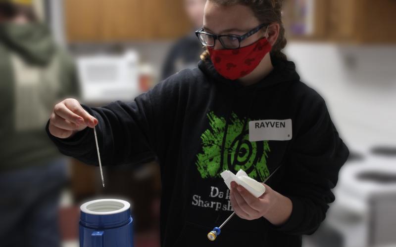 a young girl wearing a black sweater with green writing on it holding a small tube above a blue thermos