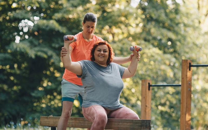 Female fitness instructor assisting an adult female at an outdoor gym.