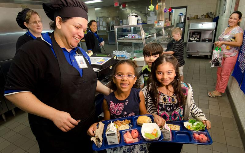 School lunch staff serving trays of healthy food a group of children.