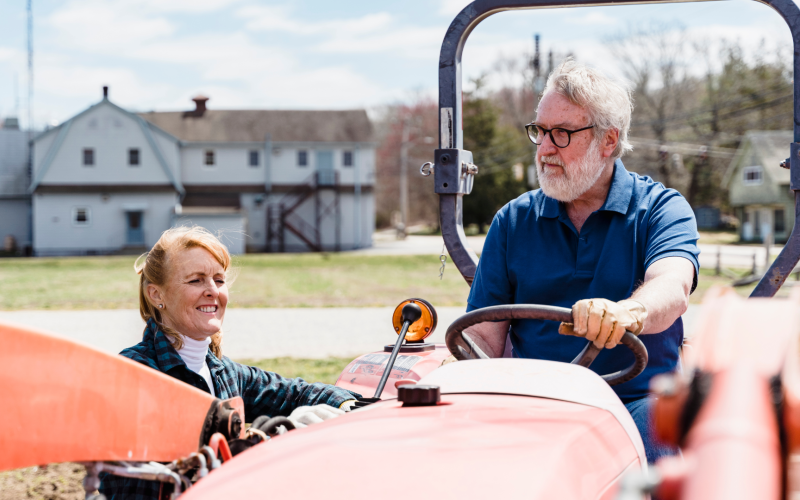 An older man sitting on a tractor with his wife standing on the ground talking to him