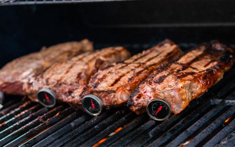Strip steaks on grill with meat thermometers properly inserted through the side of the cuts.
