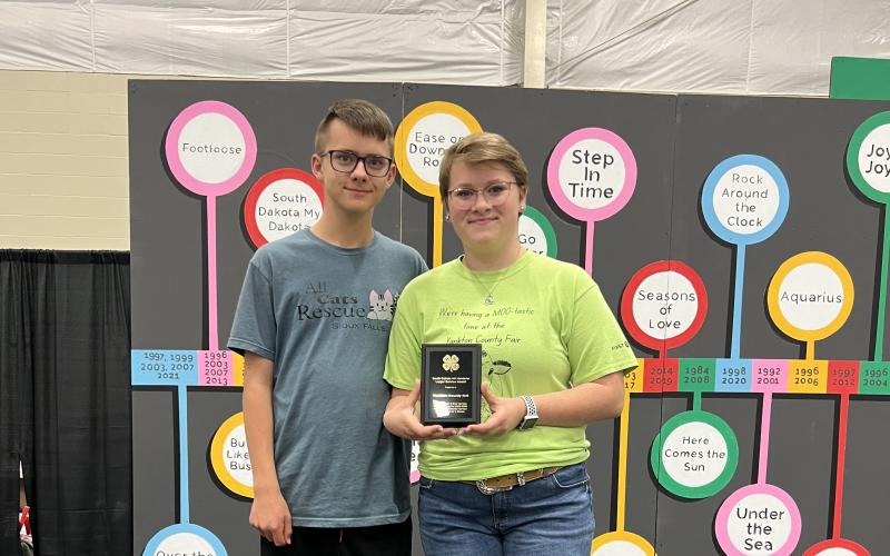 a teenage boy with short hair and glasses wearing a grey t-shirt and shorts stands next to a teenage girl with short hair and glasses wearing a bright green t-shirt and jean shorts holding an award plaque