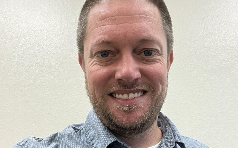a man in a blue shirt with light brown hair and a light beard smiles at the camera