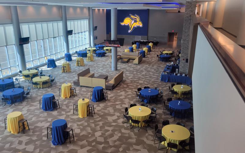 a room is pictured from above with round tables covered in blue and yellow tablecloths. a large picture of the SDSU jackrabbit logo is on the far wall