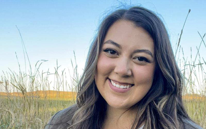 Kaylee Wheeler smiles at the camera. Her long brown hair is down, and she is standing outside in a field