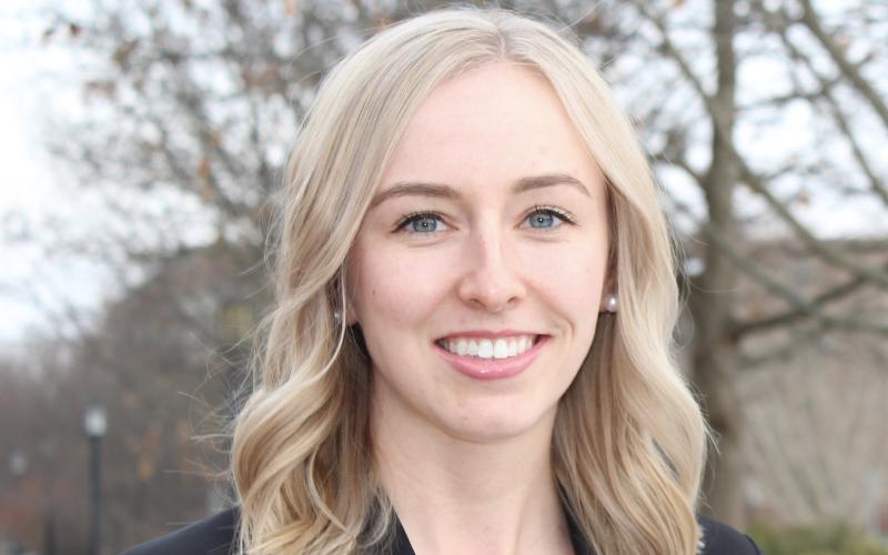 Sarah Sellars smiles at the camera. Her shoulder-length blonde hair is down, and she is wearing a purple top with a black blazer