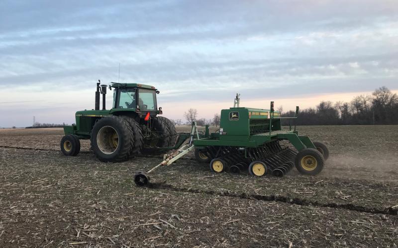 A green tractor pulls a planter to plant oats in a field
