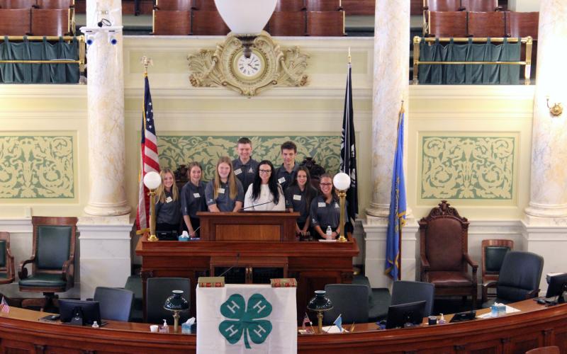 Nine youth stand behind a podium inside the South Dakota Capitol session chamber. A 4-H flag is draped in front.