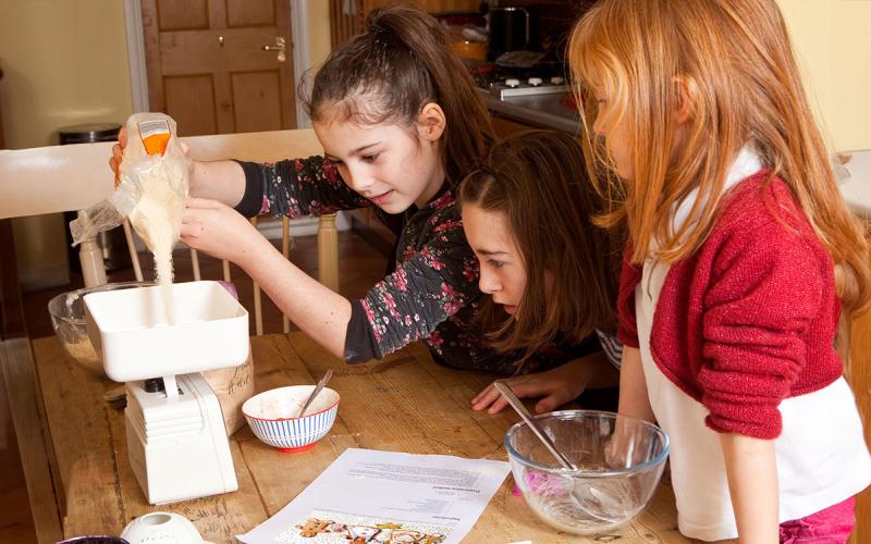 Three young girls weighing sugar on a kitchen scale.|