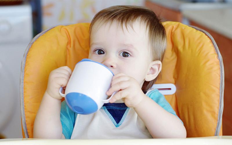 Infant boy seated in a high chair drinking from a sippy cup.