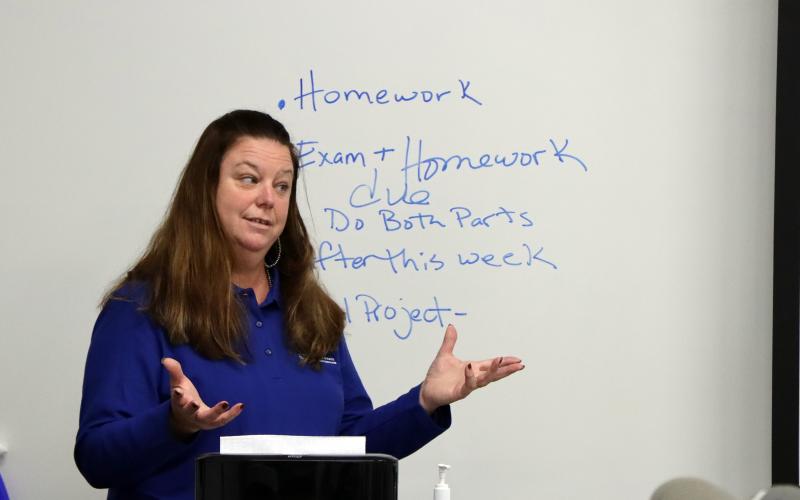 Heather Gessner gestures to her classroom. She's wearing a blue shirt and standing in front of a white board with the bullet points homework, exam plus homework due, do both parts, after this week