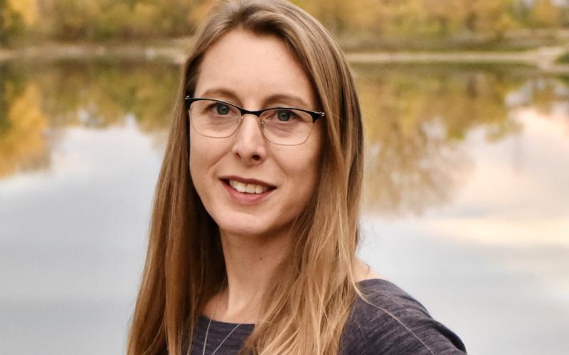 Robin Buterbaugh smiles at the camera while standing in front of a body of water with trees in the distant background