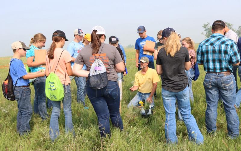 A man in a yellow shirt kneels in a pasture with a group of youth and teenagers standing in a circle around him