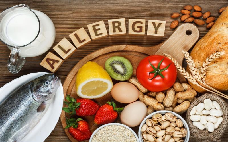 A variety of common food allergens arranged on a table surrounded by the word “Allergy” spelled out on wooden tiles.