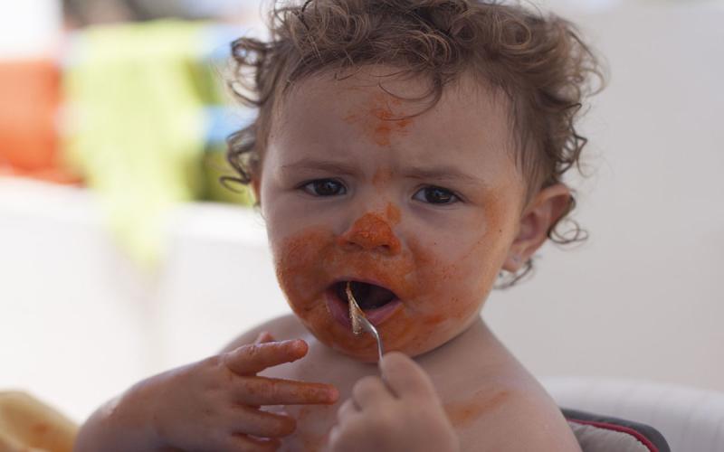 Infant grasping a training fork with tomato sauce all over her face.