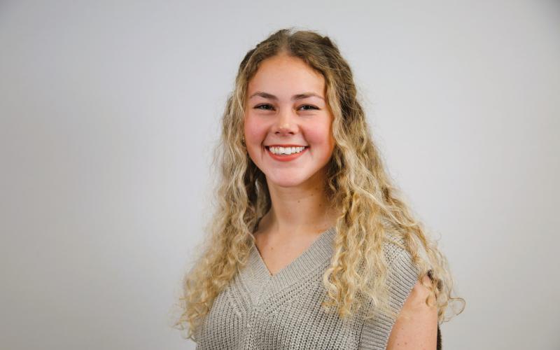 Addie Womack smiles at the camera. She has long, curly blonde hair and is wearing a grey shirt