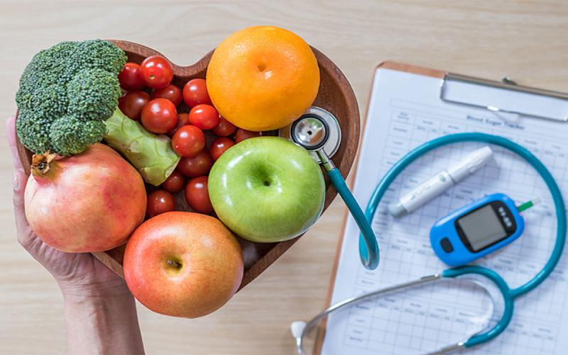 Hand holding a heart-shaped bowl of fruits and vegetables near a clipboard, stethoscope, and glucose meter.