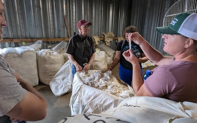 A man in a warehouse surrounded by bags of wool speaks to a group of people