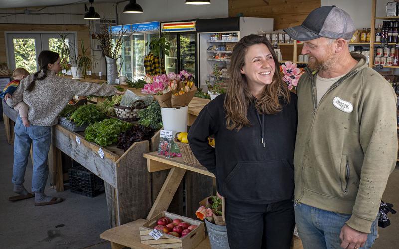 Owners of a specialty crop farm smiling at each other in a small on-farm market as a customer shops in the background.